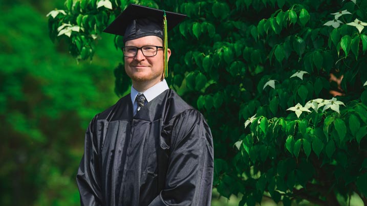 The image shows James Brindley wearing a graduation cap and gown standing outdoors in front of lush green foliage. James is dressed in a traditional black cap and gown with a white shirt and patterned tie underneath. The background is filled with vibrant green leaves, indicating a setting that is likely in a natural or garden-like environment. James is centrally positioned, and his expression is calm and composed.