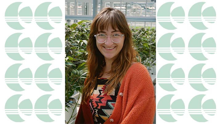 The image features a close-up of Sarah Capparuccini in a greenhouse setting. She has long, straight brown hair with bangs and is wearing clear, round glasses. She is smiling and looking directly at the camera. She is dressed in a colorful, patterned top with geometric designs in red, yellow, black, and turquoise, and an orange cardigan with visible buttons. The background shows green leafy plants in white pots, typical of a greenhouse environment, with a blurred out glass structure.