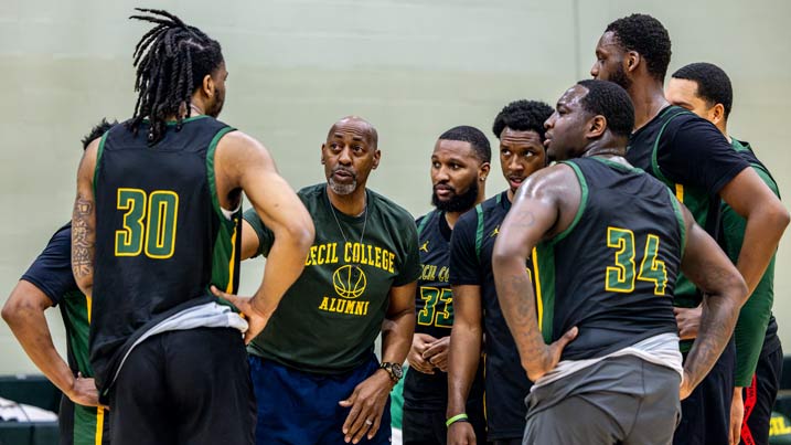 The image shows a group of seven individuals gathered indoors, in what appears to be a gymnasium. They are engaged in a discussion or strategy session, likely related to basketball. Five men are wearing black sports jerseys with green accents, displaying the numbers 30, 35, and 34 prominently. Thornton Spellman is in the center wearing a dark t-shirt with the words "Cecil College Alumni" in yellow lettering. The group is standing close together, suggesting they are listening intently and possibly strategizing. The setting includes a light-colored wall in the background.